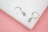 A pair of sterling silver Hamsa hand charm hoop earrings rests on a white display surface against a pink background.