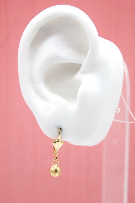 A white mannequin ear displays a delicate gold dangle earring featuring a heart charm and a textured sphere.