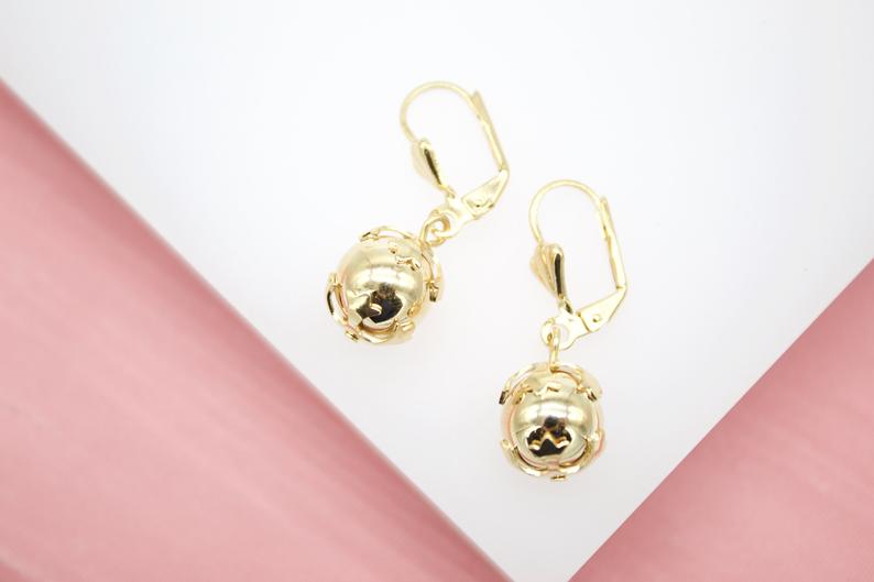 Two gold dangle earrings featuring decorative sphere charms rest on a white and pink display surface.