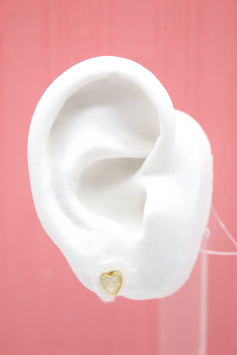 A white model ear showcases a delicate gold heart-shaped stud earring against a soft pink background.