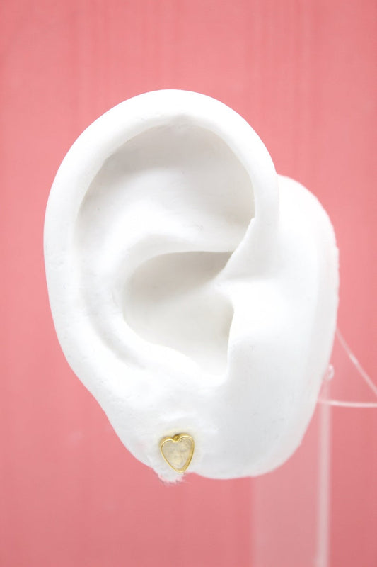 A white model ear showcases a delicate gold heart-shaped stud earring against a soft pink background.
