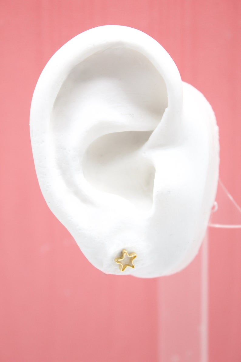 A white mannequin ear displays a small gold star stud earring against a pink background.