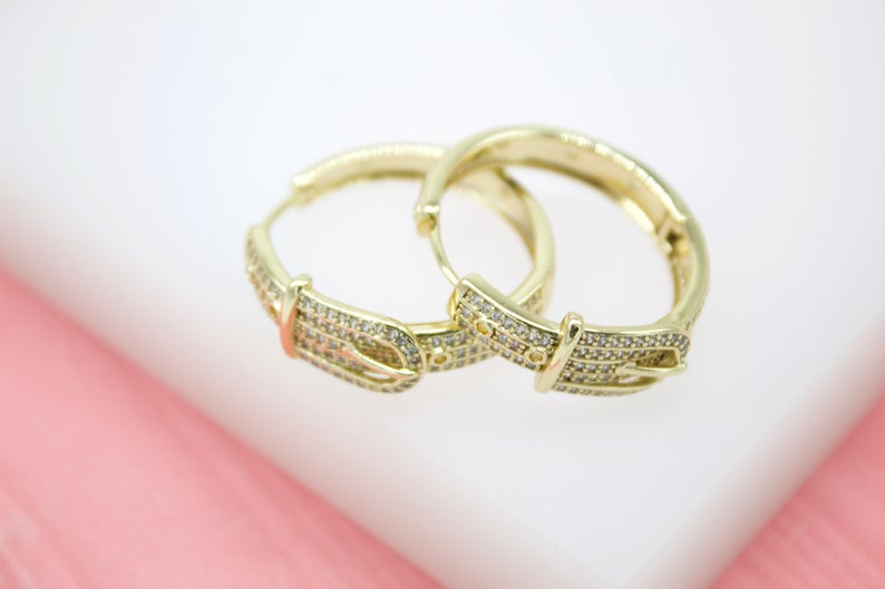 A pair of gold hoop earrings features a sparkling pave buckle design resting on a white display surface.