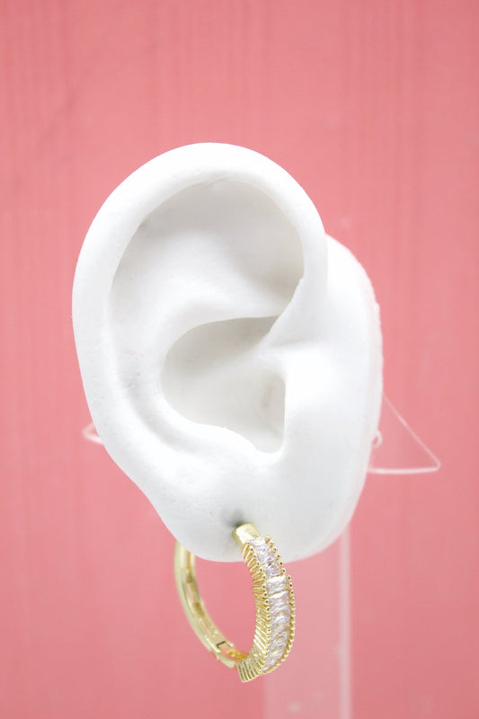 A white mannequin ear displays a gold hoop earring embedded with sparkling white baguette-cut stones against a pink background.