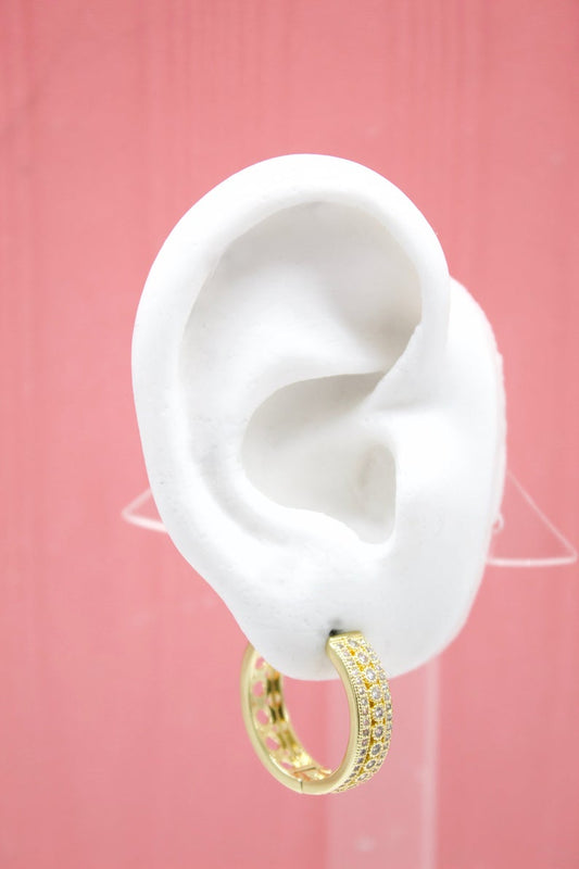 A white display ear model showcases a sparkling gold pave huggie hoop earring against a soft pink background.