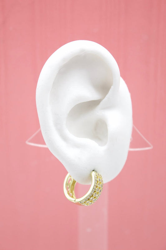 A white mannequin ear displays a sparkling gold huggie hoop earring encrusted with tiny cubic zirconia stones against a pink background.