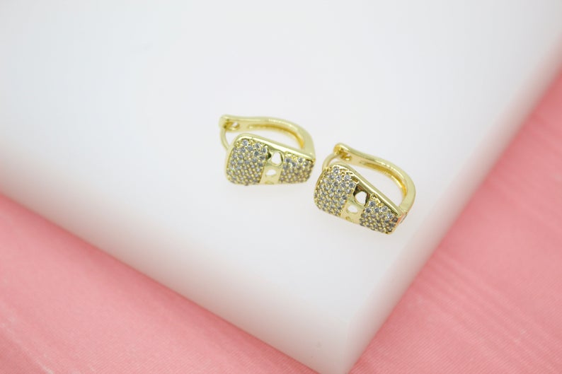 A pair of gold huggie hoop earrings encrusted with sparkling cubic zirconia stones rests on a white display surface.
