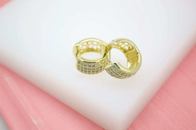 A pair of sparkling gold huggie hoop earrings encrusted with tiny cubic zirconia stones rests against a soft white and pink background.