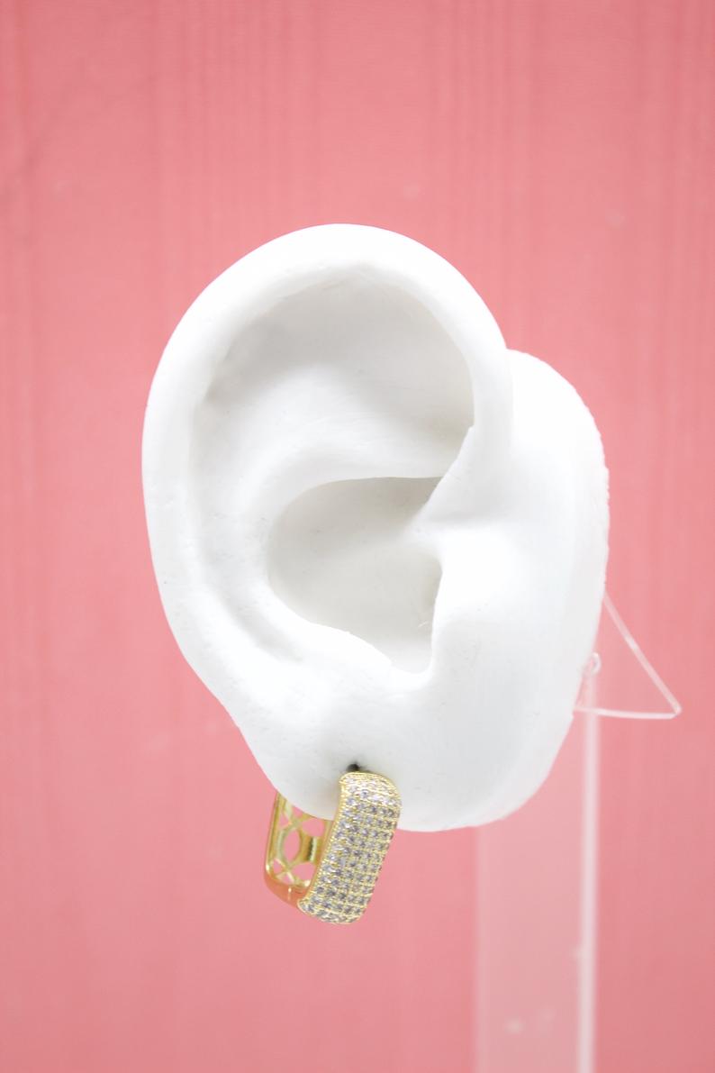 A white mannequin ear displays a gold huggie hoop earring encrusted with sparkling cubic zirconia stones against a pink background.
