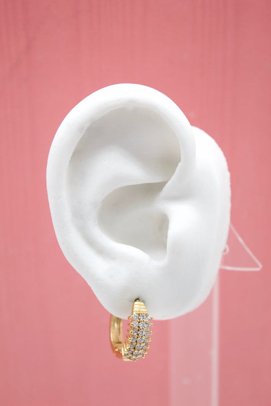 A white mannequin ear displays a sparkly gold pave huggie hoop earring against a soft pink background.