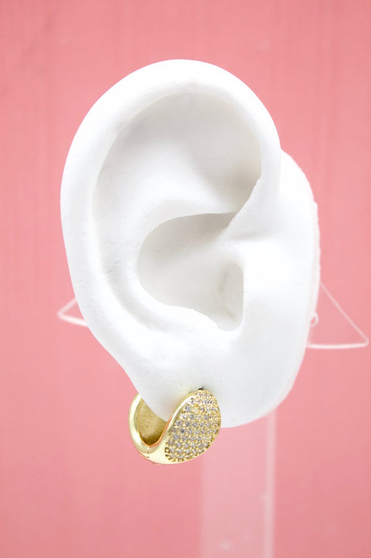 A white mannequin ear displays a chunky gold huggie hoop earring encrusted with sparkling pave cubic zirconia stones against a soft pink background.