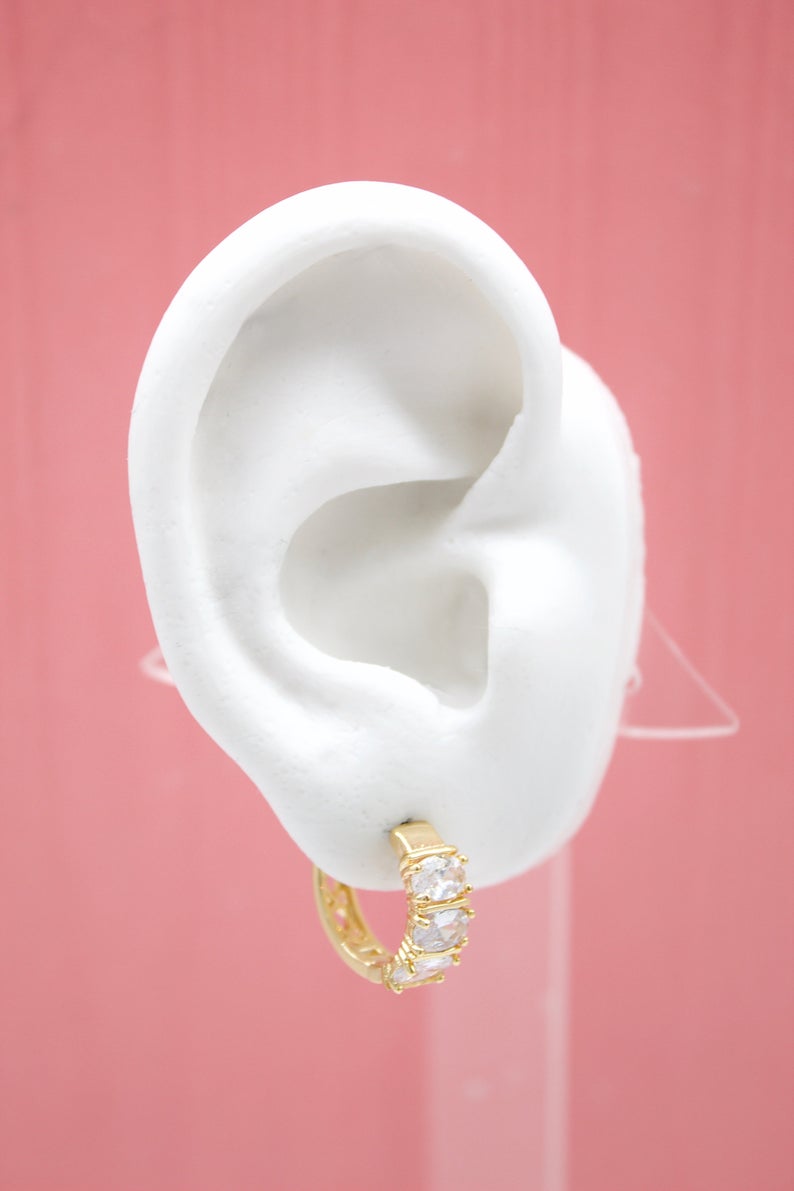 A white mannequin ear displays a gold hoop earring sparkling with inlaid cubic zirconia stones against a pink background.