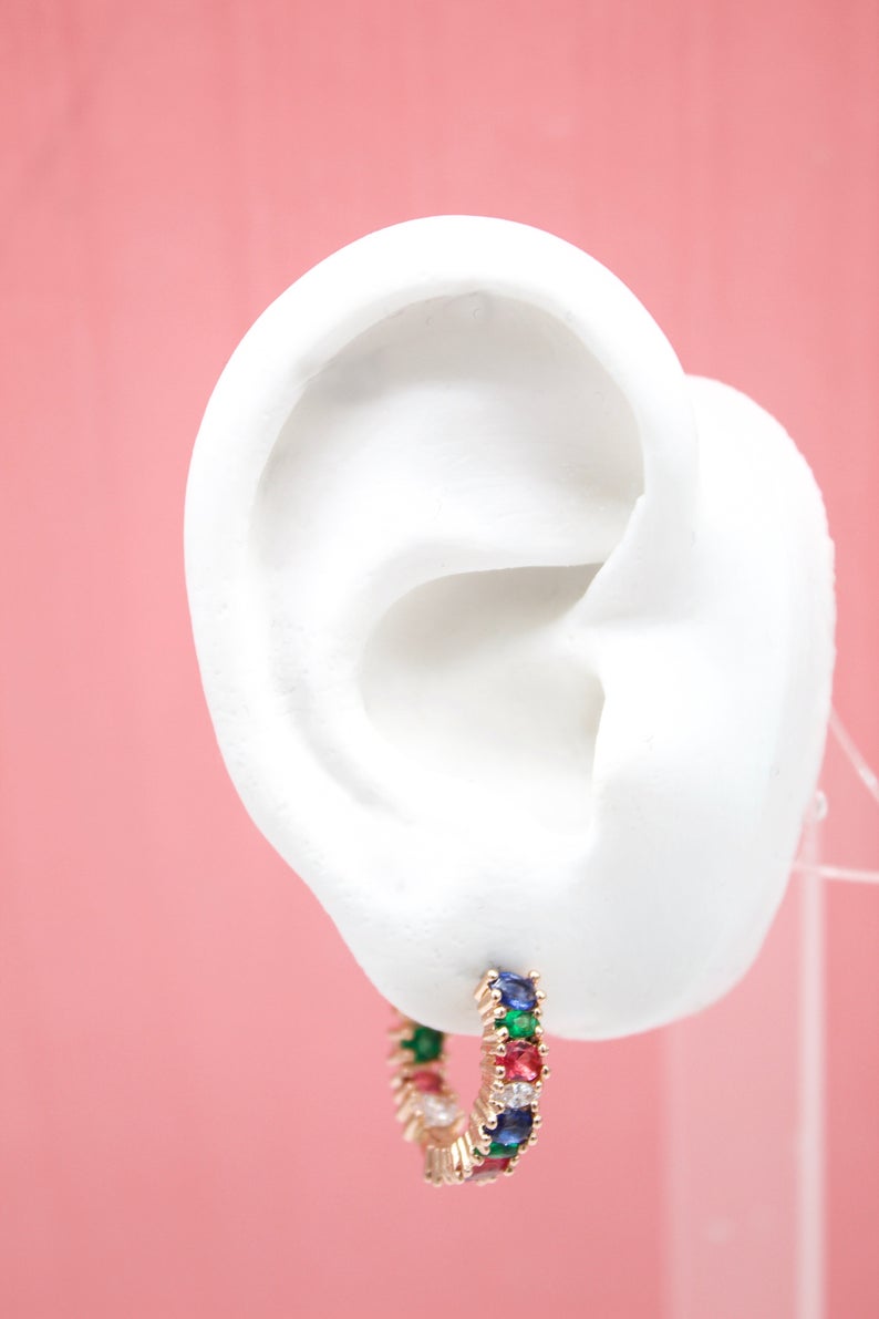 A white mannequin ear displays a vibrant rainbow huggie hoop earring featuring multi-colored gemstones against a pink background.