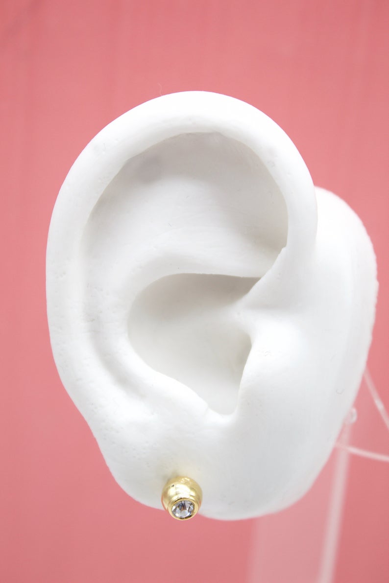 A white model ear displays a small gold stud earring with a sparkling gemstone against a pink background.