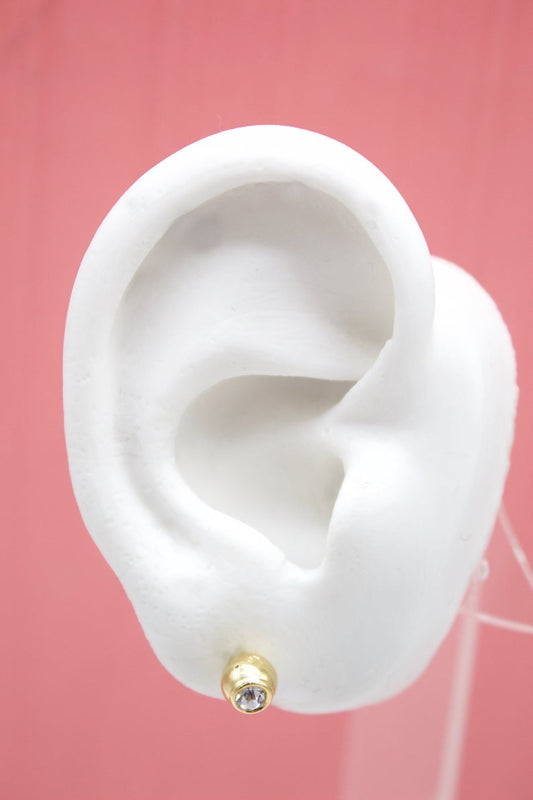 A white model ear displays a small gold stud earring with a sparkling gemstone against a pink background.