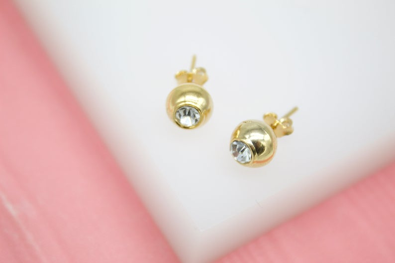 A pair of gold ball stud earrings featuring embedded clear crystals rest on a white display surface.