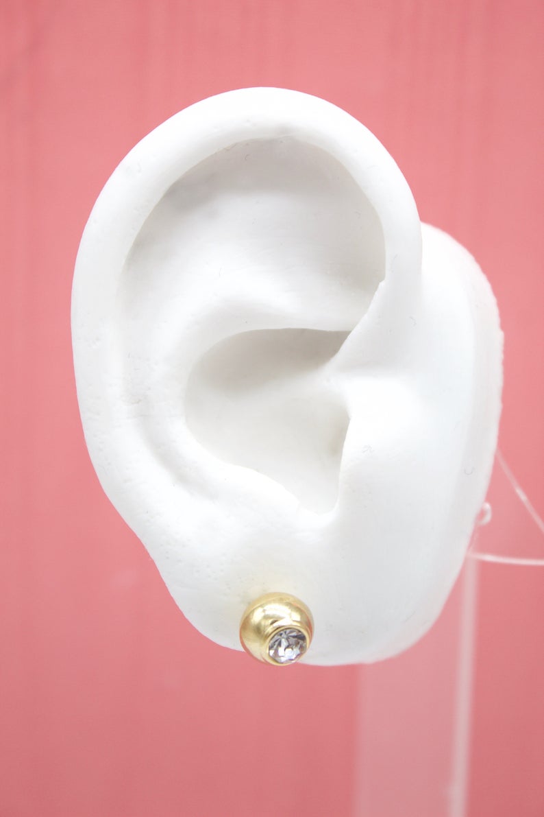 A white mannequin ear displays a shiny gold ball stud earring with a central diamond rhinestone against a pink background.