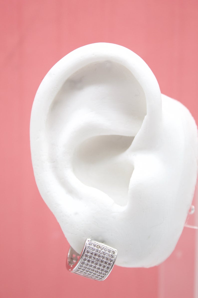 A white model ear displays a silver pave huggie hoop earring against a pink background.