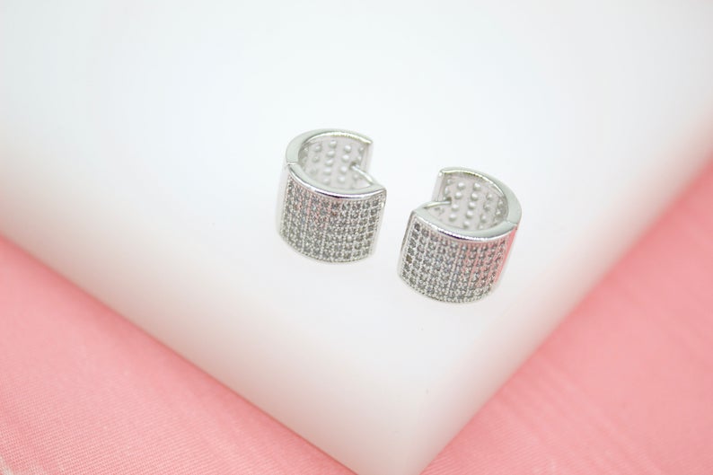 A pair of silver pave huggie hoop earrings rests on a white and pink display surface.
