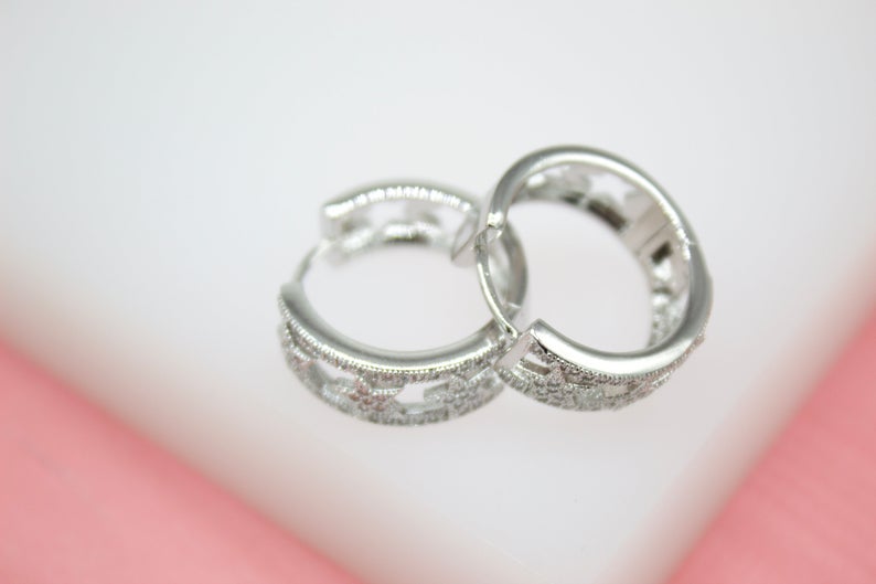 Two silver hoop earrings rest on a white surface, showcasing intricate cut-out patterns and sparkling crystal embellishments.