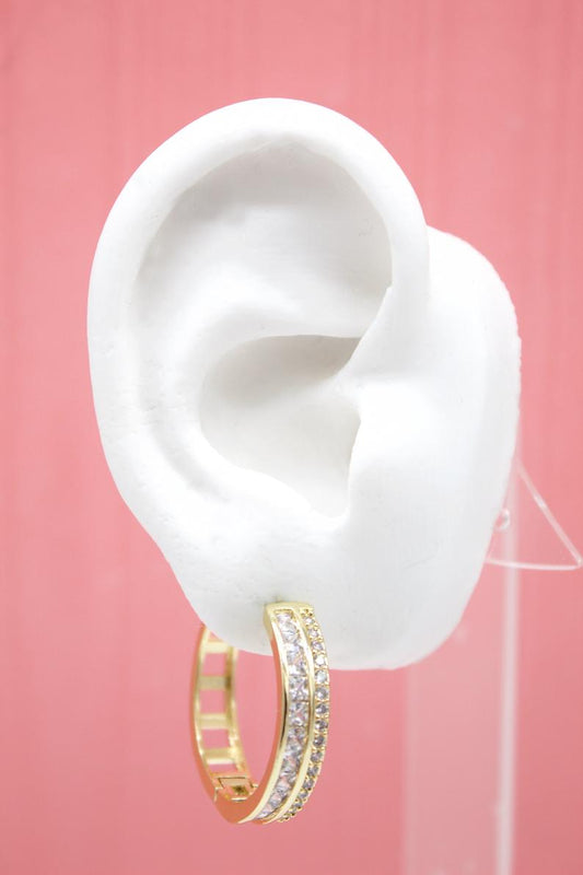 A white mannequin ear displays a glittering gold hoop earring encrusted with clear cubic zirconia stones against a pink background.