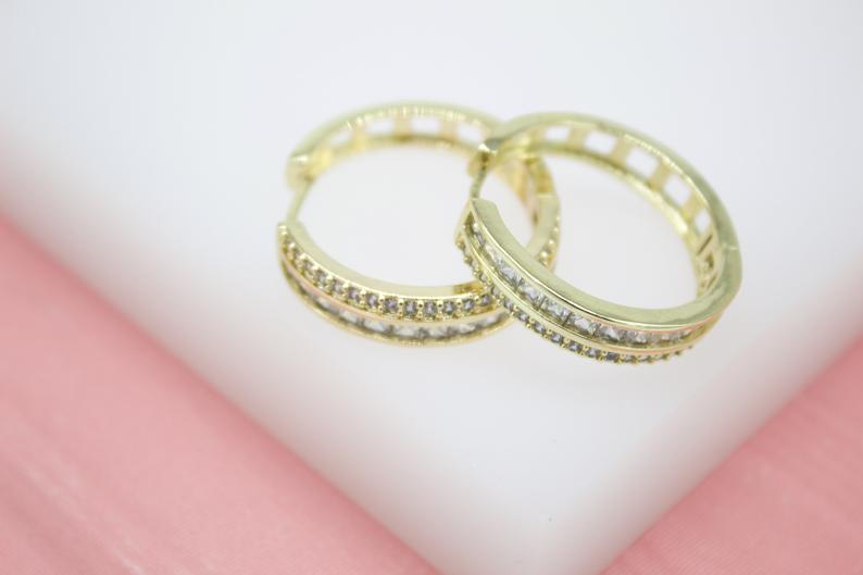A pair of gold hoop earrings embellished with sparkling cubic zirconia crystals rests on a white and pink surface.