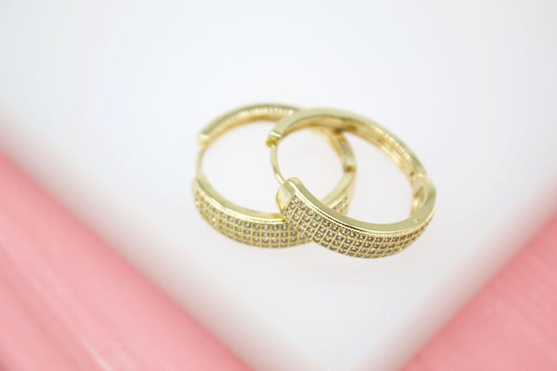 A pair of pave-set diamond gold hoop earrings rests on a white display surface against a pink background.