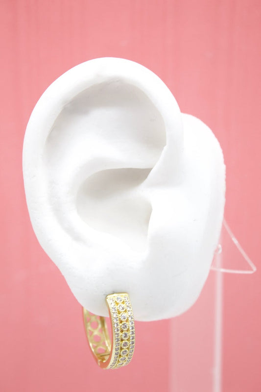 A white mannequin ear displays a sparkling gold huggie hoop earring encrusted with small clear crystals against a pink background.