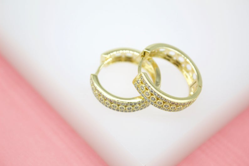 Two gold pave huggie hoop earrings rest on a white and pink display surface.