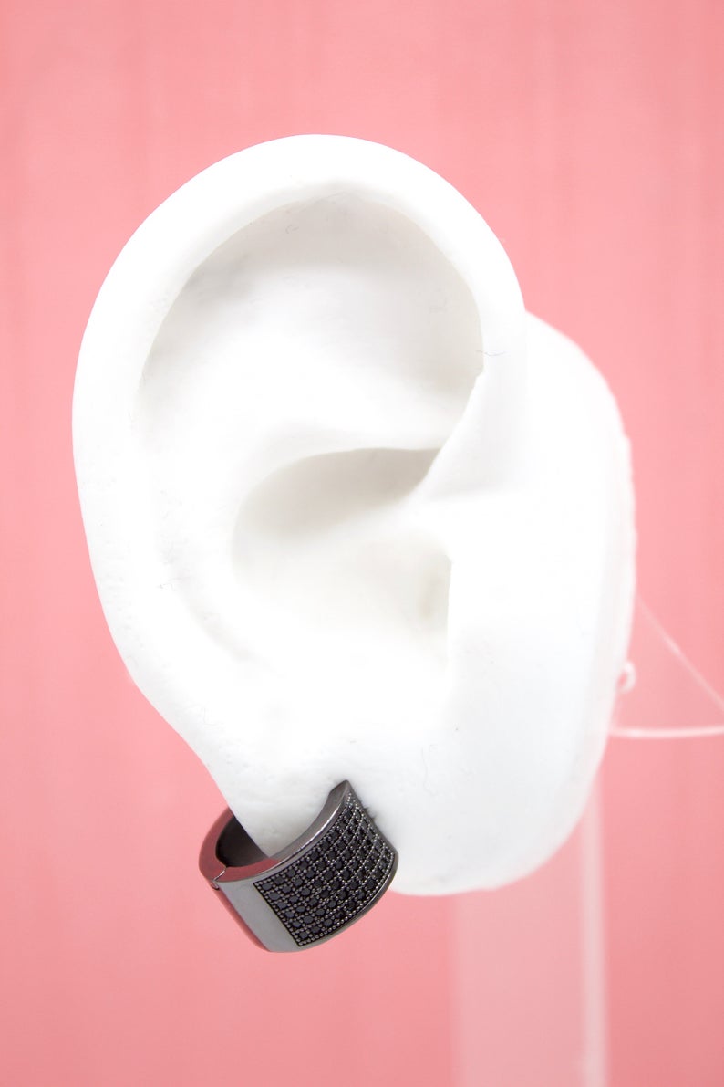 A white mannequin ear displays a wide, black pave huggie hoop earring against a soft pink background.