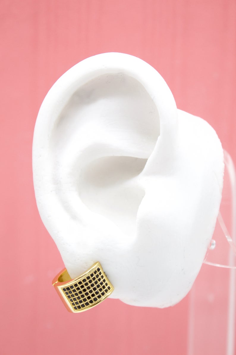 A white mannequin ear displays a chunky gold huggie hoop earring encrusted with black pave stones against a pink background.