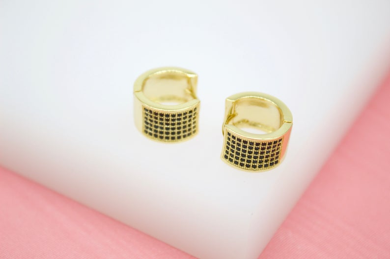 A pair of gold huggie hoop earrings featuring intricate black pave stones rest on a white surface.