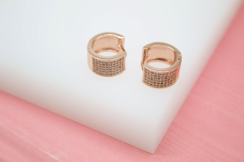 Two rose gold huggie hoop earrings with paved cubic zirconia stones rest on a white display block.