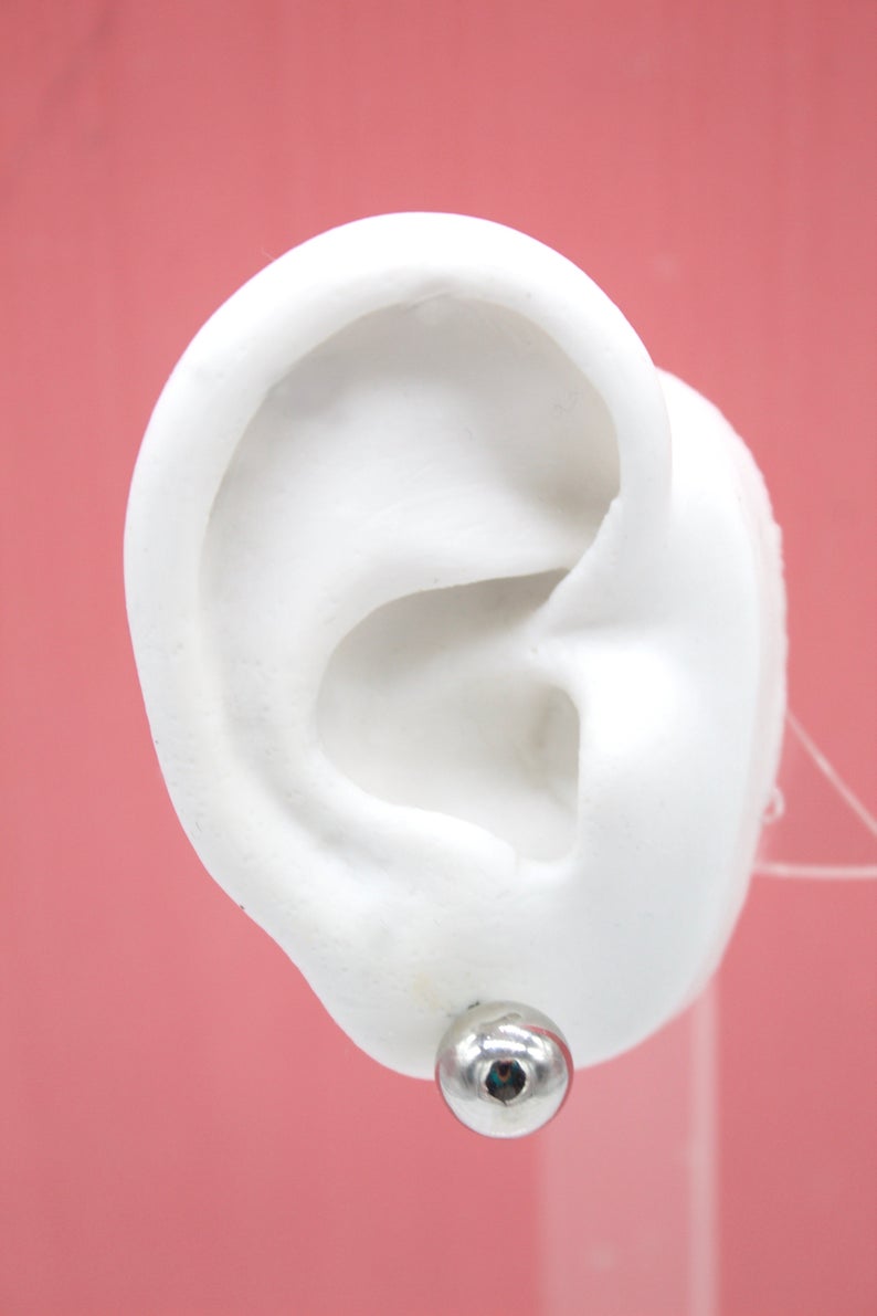 A white ear model displays a shiny silver ball stud earring fastened to the lobe against a solid pink background.