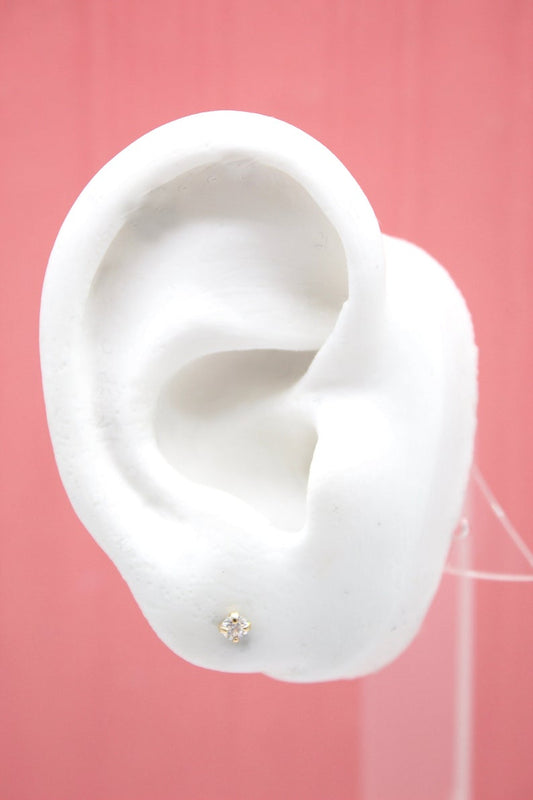 A small gold diamond stud earring sparkles on the lobe of a white mannequin ear against a solid pink backdrop.