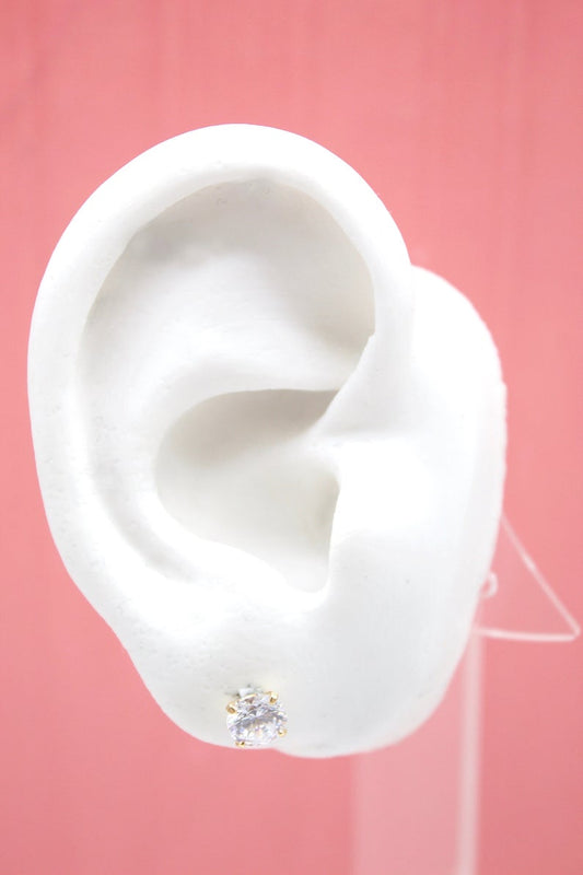 A white mannequin ear displays a sparkling cubic zirconia stud earring against a soft pink background.