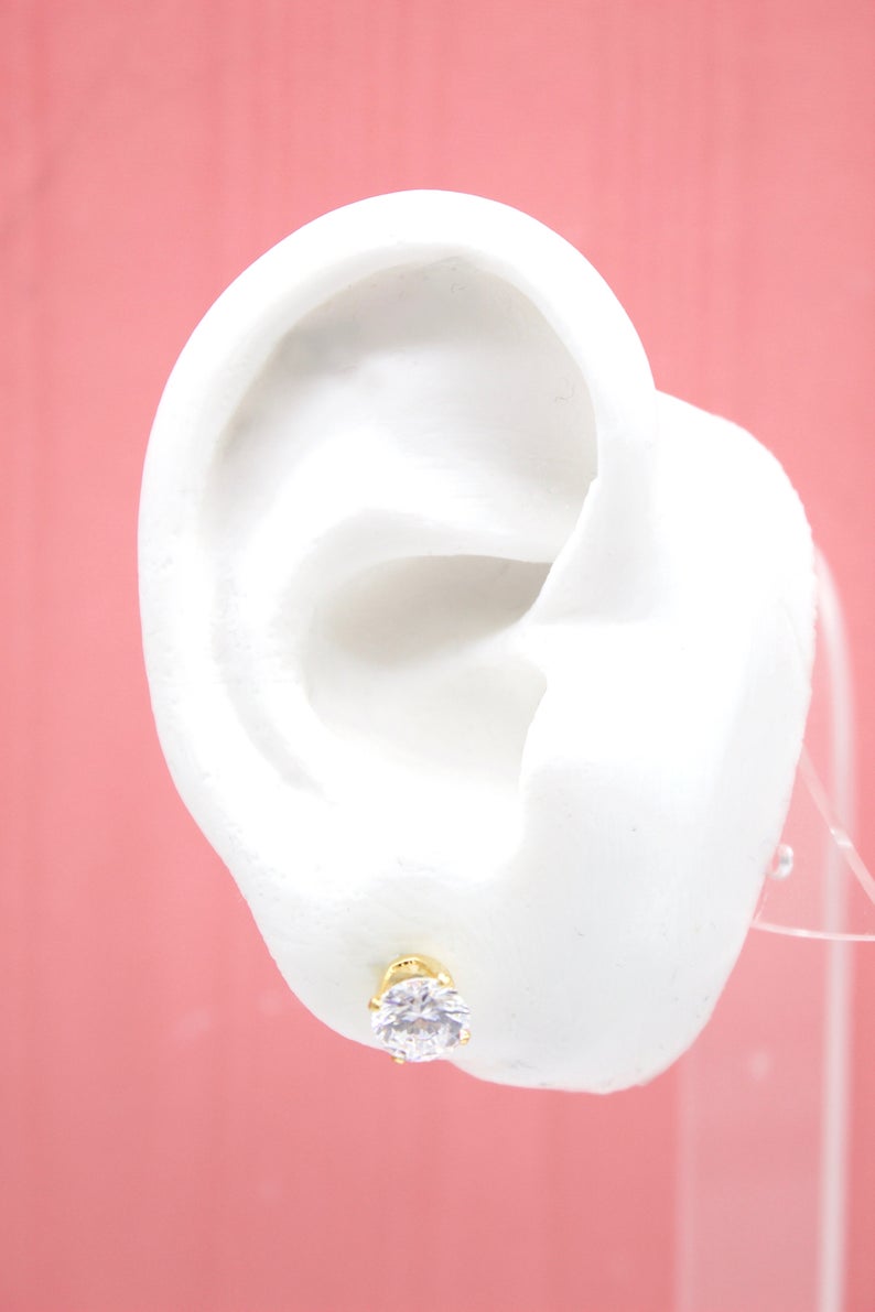 A white mannequin ear displays a sparkling gold stud earring with a brilliant round solitaire cubic zirconia against a pink background.