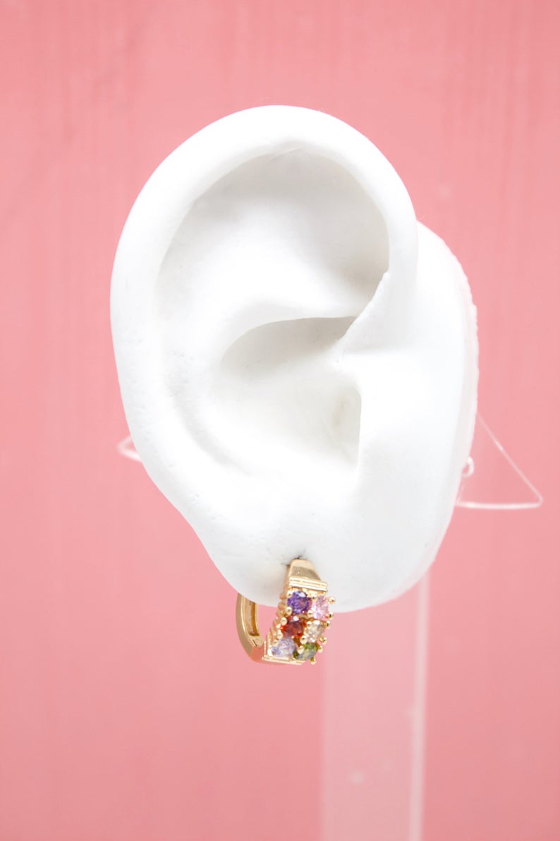 A white mannequin ear displays a gold huggie hoop earring encrusted with multicolored cubic zirconia stones against a pink background.