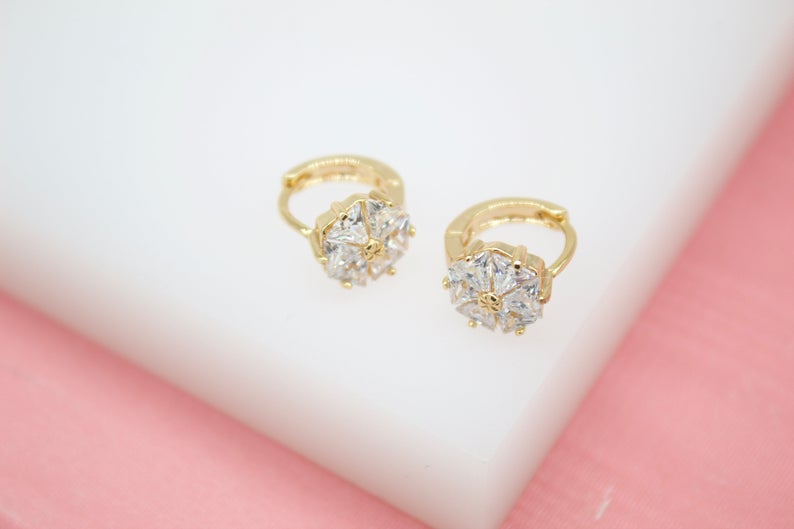 Two gold hoop earrings featuring sparkling flower-shaped zirconia crystal charms rest on a white surface.