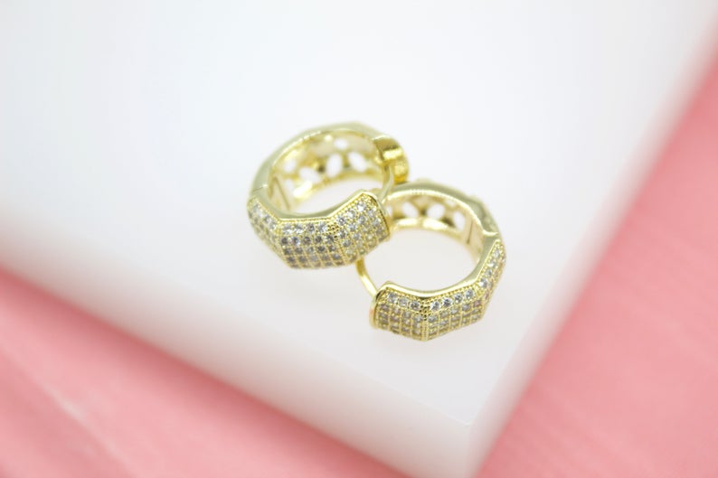 A pair of pave-set gold huggie hoop earrings rests on a white display block against a soft pink background.