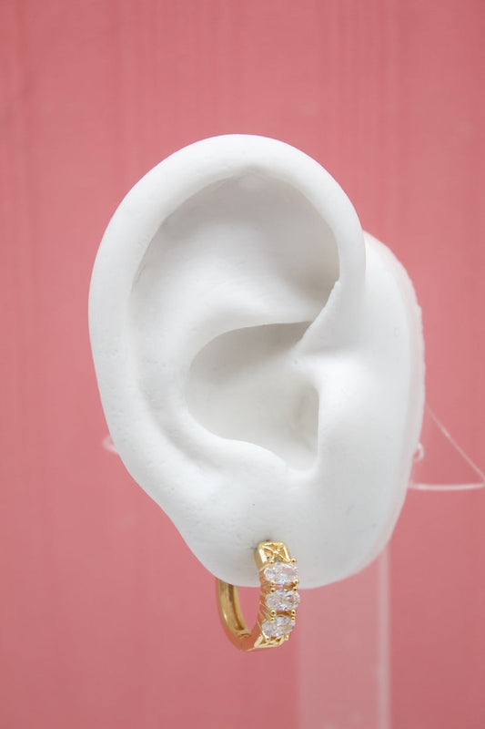 A white mannequin ear displays a gold huggie hoop earring featuring three sparkling cubic zirconia stones.