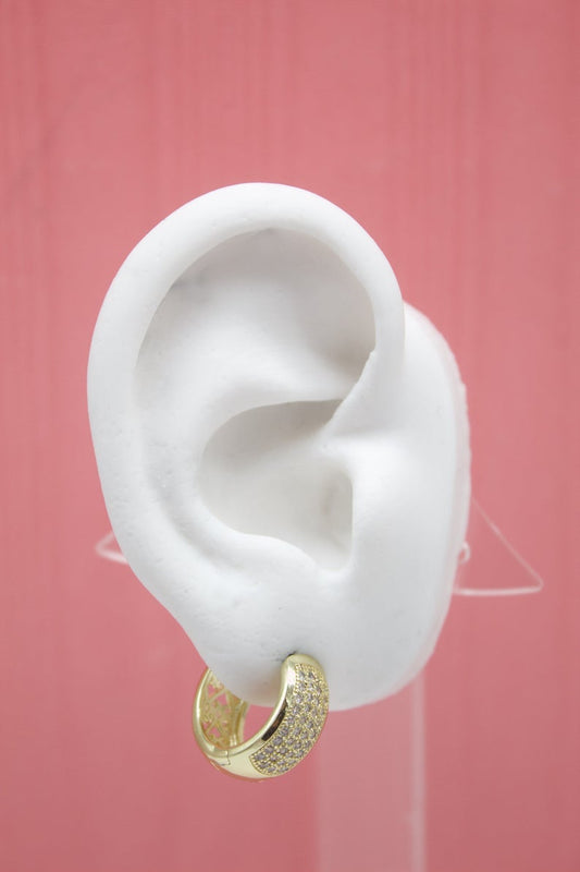 A white model ear displays a chunky gold huggie hoop earring encrusted with sparkling pave diamonds against a solid pink background.