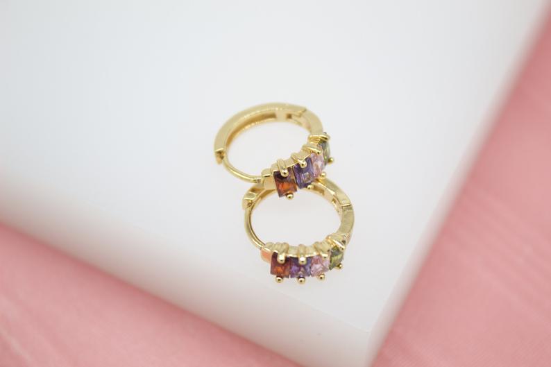 A pair of gold huggie hoop earrings features a row of colorful baguette-cut cubic zirconia stones on a white display surface.
