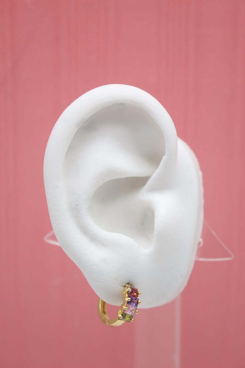 A white mannequin ear displays a gold huggie hoop earring adorned with multicolored baguette-cut stones against a pink background.