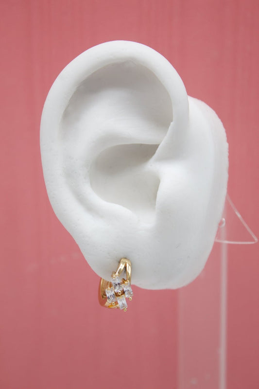 A white mannequin ear displays a sparkling gold huggie hoop earring adorned with baguette zirconia stones against a pink background.