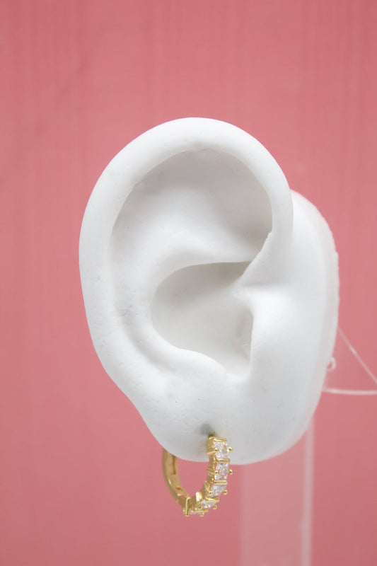 A white mannequin ear displays a sparkling gold huggie hoop earring encrusted with clear cubic zirconia stones against a pink background.