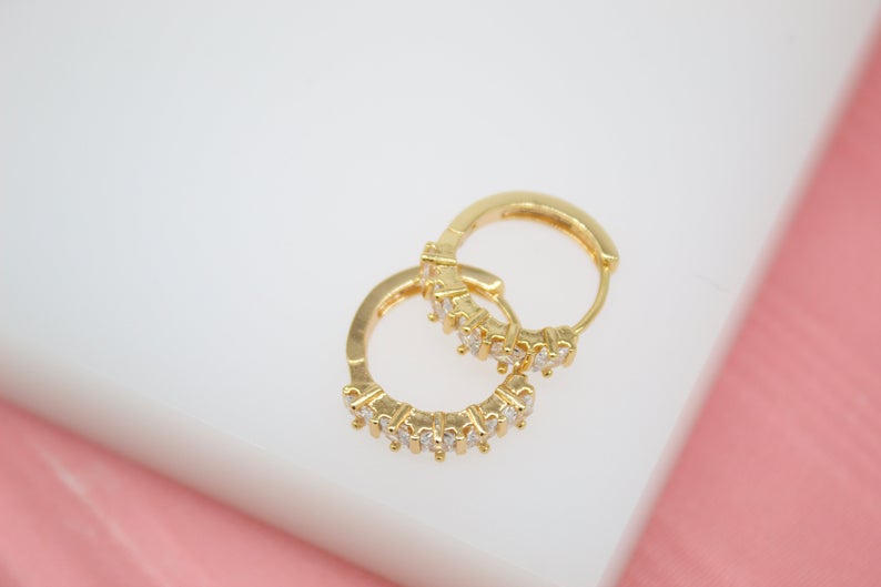 Two gold huggie hoop earrings featuring sparkling clear cubic zirconia stones rest on a white surface with a pink background.