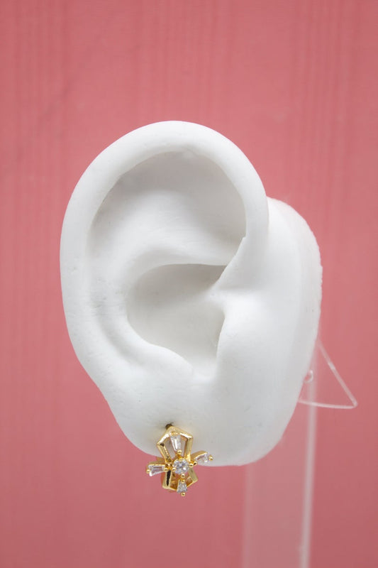 A white mannequin ear displays a gold geometric stud earring embellished with sparkling cubic zirconia crystals against a solid pink background.