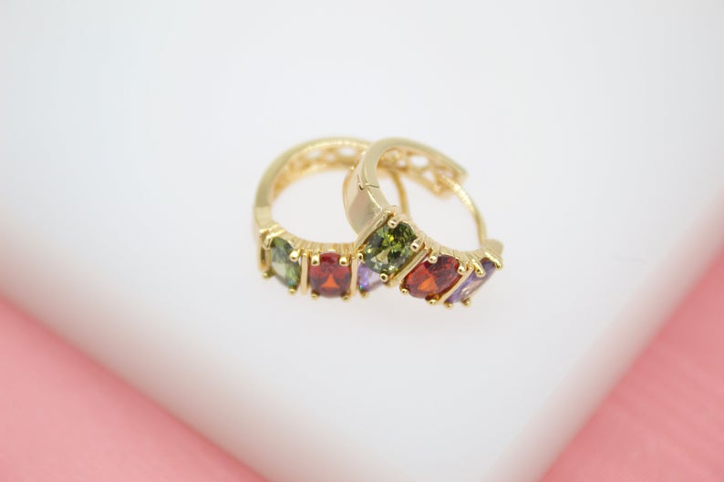 A pair of gold huggie hoop earrings features colorful multi-gemstones resting on a clean white and pink surface.