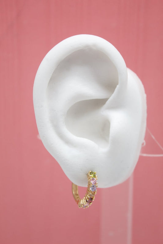 A white mannequin ear displays a gold hoop earring sparkling with colorful gemstone crystals against a pink background.